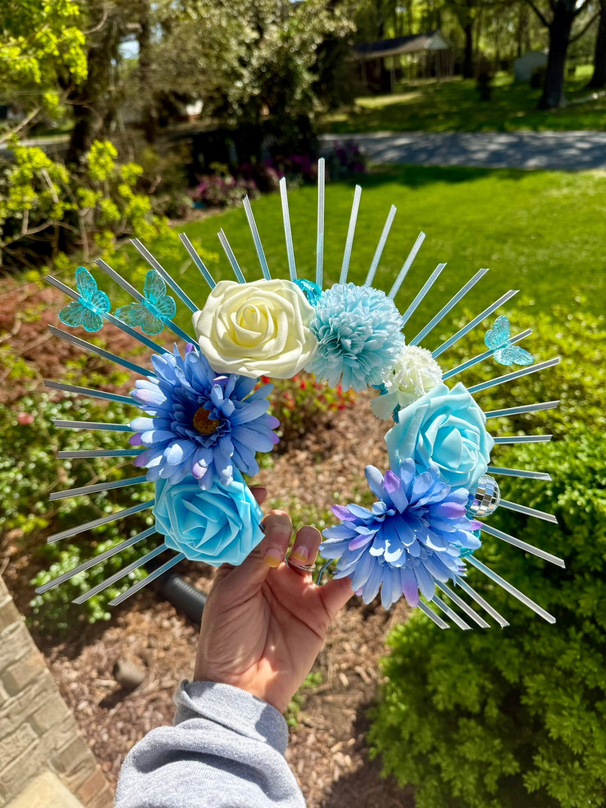 A hand holds a decorative sunburst-style flower crown outdoors in a garden. The crown has clear plastic spikes with blue, light blue, cream, and voilet artificial flowers, including roses and daisies, plus small blue butterflies.