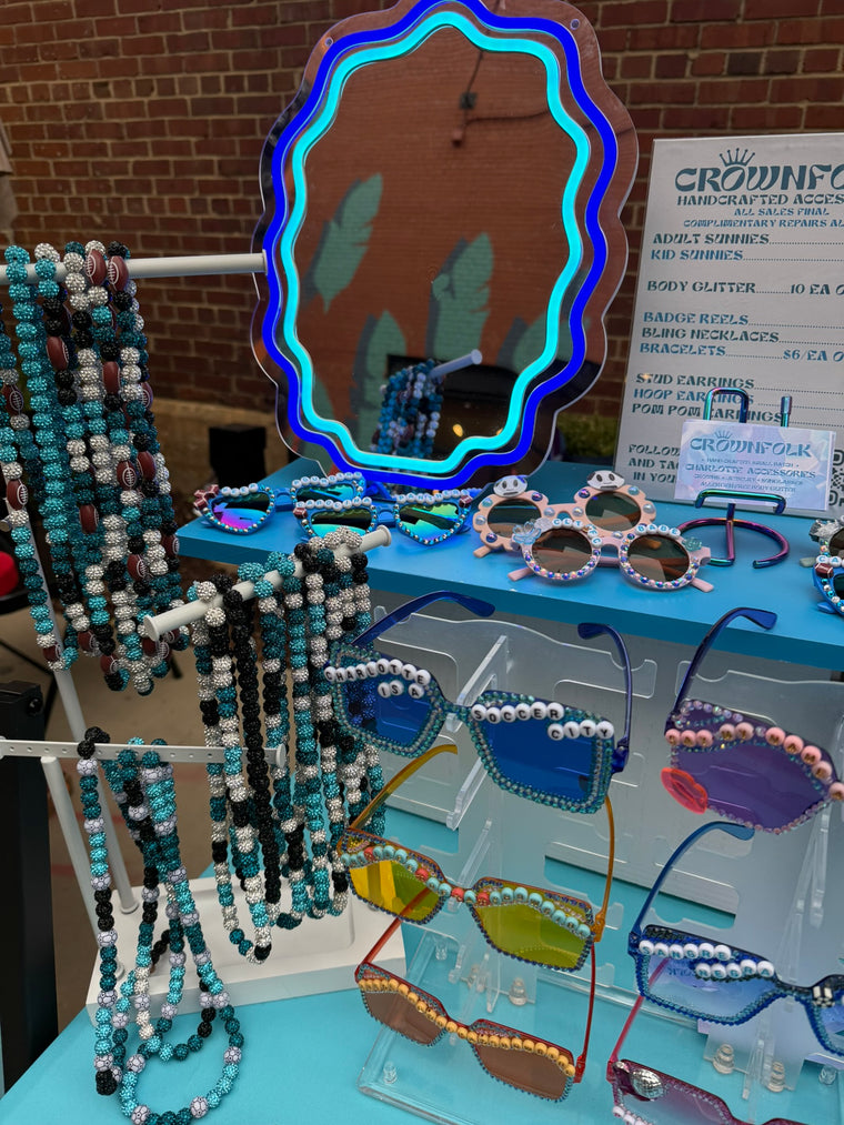 A display of novelty beaded sunglasses and matching beaded necklaces on clear stands, with a large decorative mirror and a sign reading CROWFOLK HANDCRAFTED ACCESSORIES in the background.