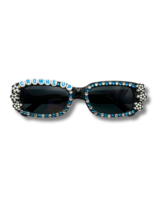 A pair of black sunglasses embellished with blue rhinestones and silver rhinestones, featuring the word 'CROWNS UP' spelled out in rhinestone letters across the top frame.