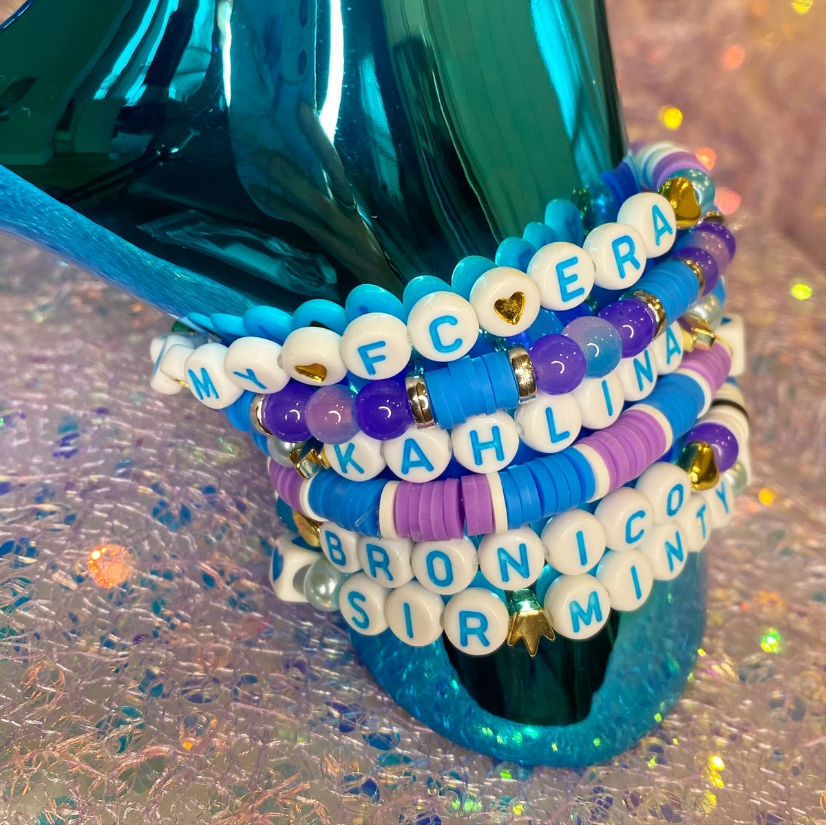 A stack of five beaded bracelets displayed on a shiny teal stand. The bracelets feature white letter beads spelling out names and phrases including 'MY FCERA', 'KAHLINA', 'BRONCO SIR', and 'MINT'.
