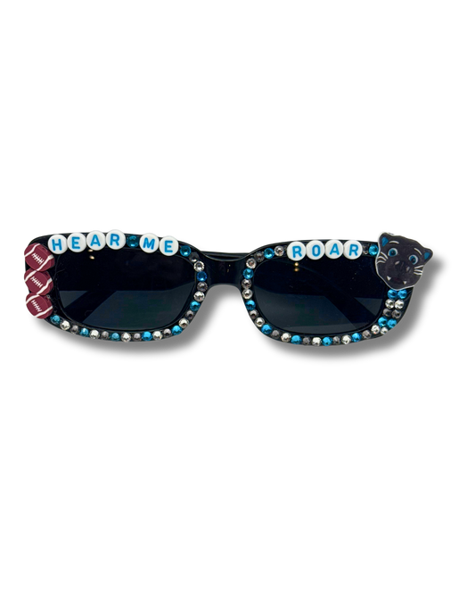 Black cat-eye sunglasses embellished with rhinestones, blue and white lettering spelling 'HEAR ME' and 'ROAR', a football accent side detail, and a black cat face on the other side.