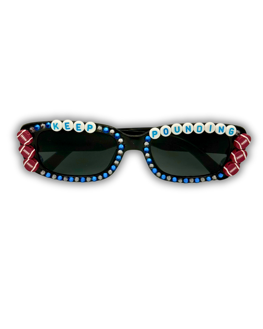 Black sunglasses with blue rhinestone trim, red-and-white striped side accents, and white letter beads spelling 'KEEP POUNDING' across the top of the lenses, shown on a plain white background.