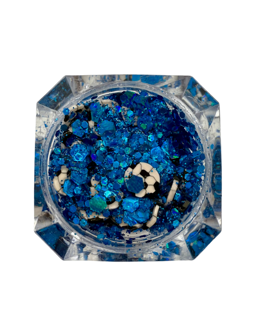 Top-down view of a clear faceted octagonal container filled with blue, navy, and black glitter flakes, set against a plain white background.
