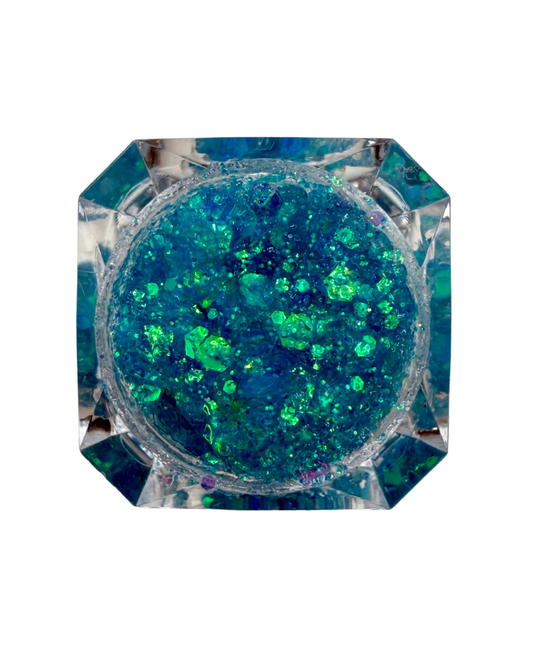 Top-down view of a clear faceted octagonal container filled with blue, green, and teal glitter flakes, set against a plain white background.