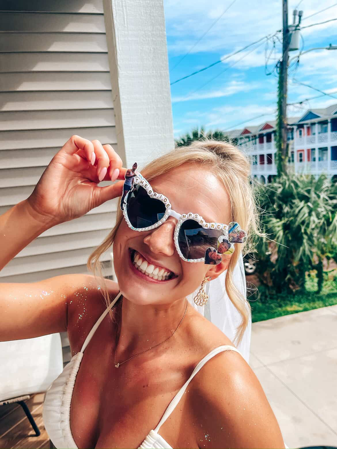 A smiling blonde woman outdoors wearing heart-shaped sunglasses with rhinestone trim and colorful mirrored lenses, along with a white bikini top and a veil.