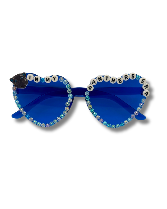 Blue heart-shaped sunglasses with rhinestone trim, letter tiles spelling 'IN MY PANTHERS ERA' on the upper rims, and a black panther charm on the left side, shown against a plain white background.