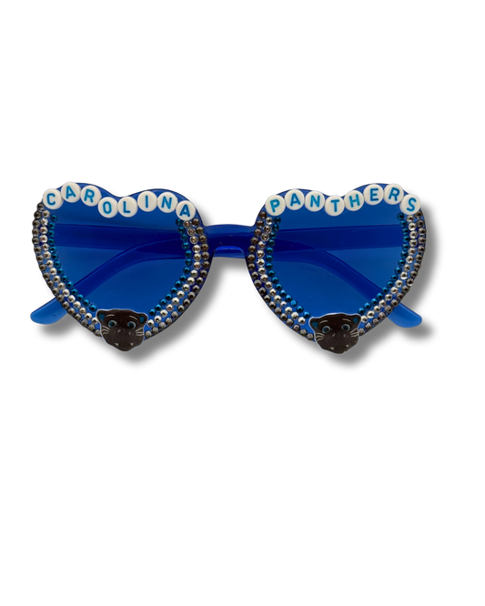 Heart-shaped sunglasses with blue mirrored lenses, rhinestone-studded borders, white letter tiles spelling 'CAROLINA PANTHERS' on top, and black panther head charms near the lower corners.