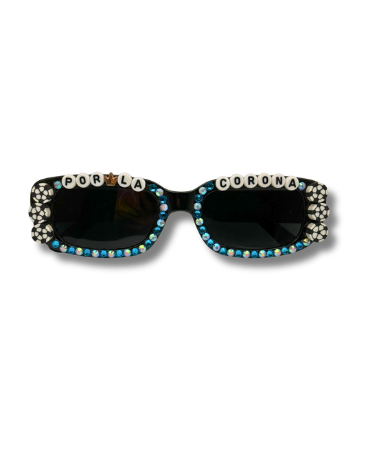 Black rectangular sunglasses embellished with rhinestones and letter beads spelling 'POR LA' on one side and 'CORONA' on the other, with blue and white rhinestones along the lower frame and soccer ball accents on both sides of the frame, shown on a white background.