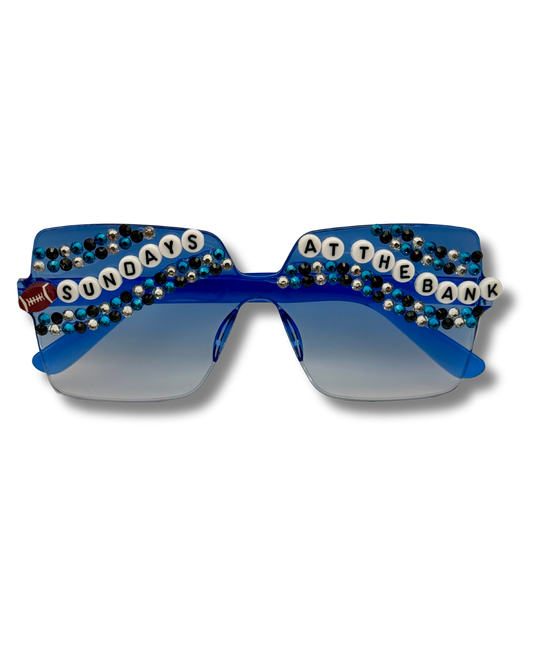 Blue novelty sunglasses decorated with rhinestones, beaded letter tiles spelling 'SUNDAYS AT THE BANK', and a small football on the left temple, shown on a white background.